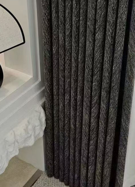 a close-up view of a dark grey curtain with a herringbone pattern, hanging next to a white wall.