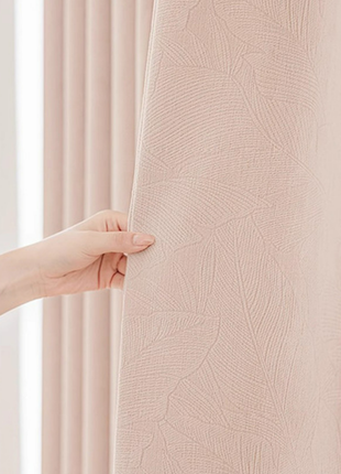 A hand is holding a curtain with a leaf pattern, partially covering a window.